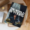 A dark poster titled The World Is Yours featuring a man in a suit and a gold briefcase, displayed on a light wooden surface.
