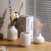 A set of three minimalist white ceramic decorative vases with dried floral arrangements on a wooden surface.