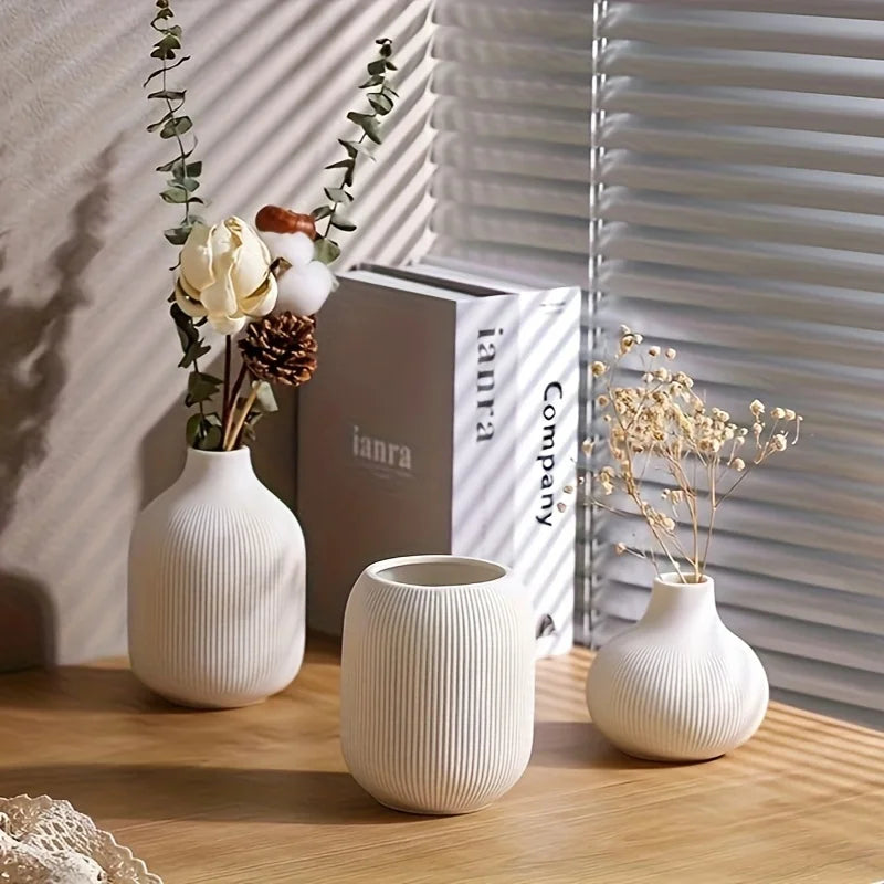 A set of three minimalist white ceramic decorative vases with dried floral arrangements on a wooden surface.