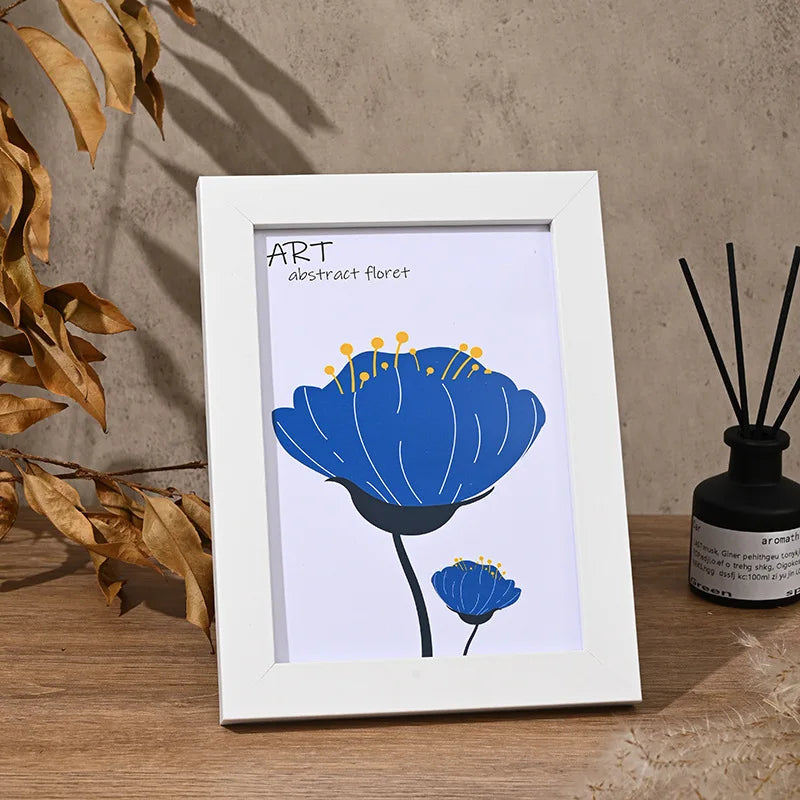A white frame holds an abstract blue flower illustration on a piece of paper, displayed on a wooden surface.
