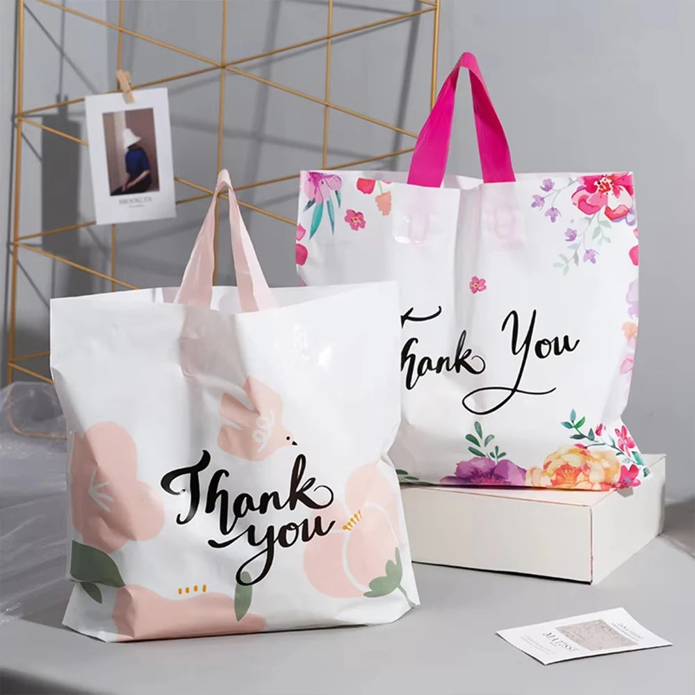 White floral thank you gift bags with pink handles displayed on a light gray surface.