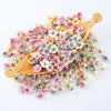 A collection of silk artificial flower heads in shades of pink, blue, and cream are displayed on a wooden paddle.