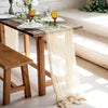 Sage green gauze table runner draped over a wooden table with glassware and greenery.