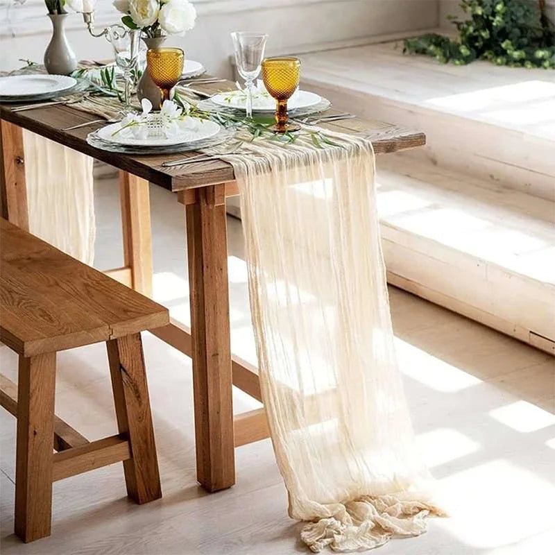 Sage green gauze table runner draped over a wooden table with glassware and greenery.