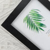 A black photo frame displaying a watercolor illustration of green palm leaves on a white background.