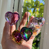 A hand holds several iridescent, heart-shaped glass pendants hanging by thin strings against a bright window background.