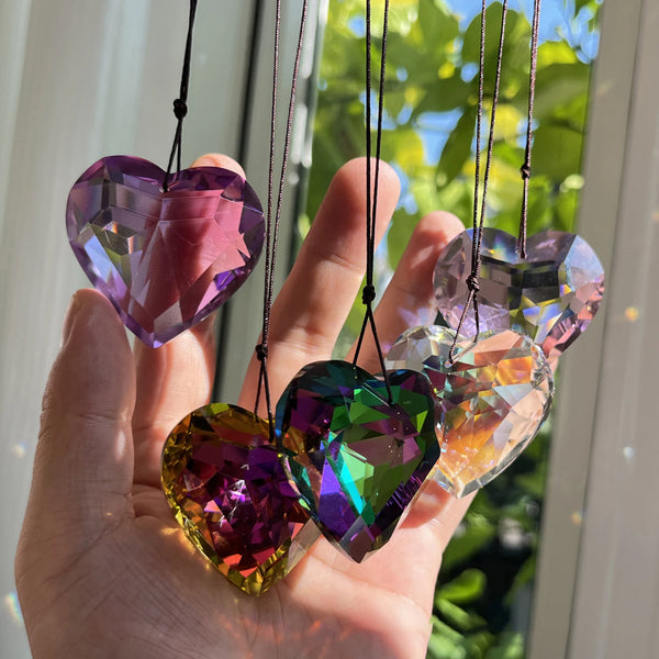 A hand holds several iridescent, heart-shaped glass pendants hanging by thin strings against a bright window background.
