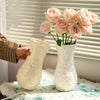 A white ceramic vase with a relief pattern is displayed on a table next to a hand holding it, set against a window blind.