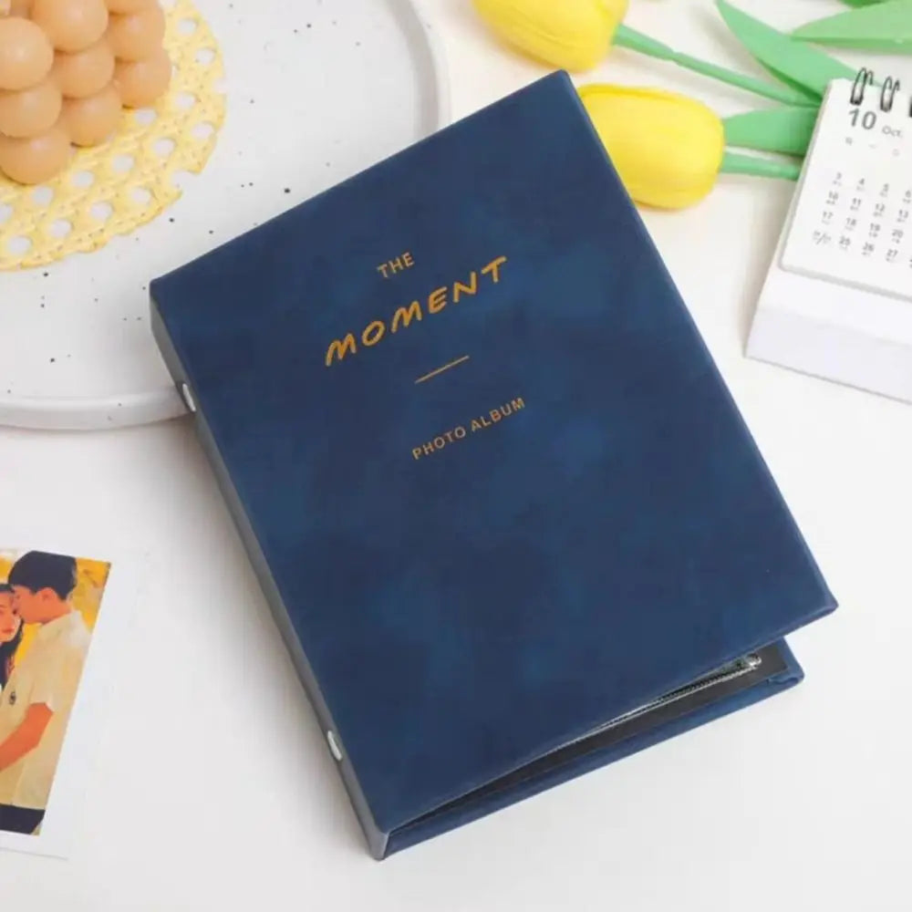 A dark blue photo album titled The Moment with gold lettering resting on a white surface next to yellow flowers and a white c