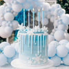 A light blue frosted birthday cake decorated with white icing and lit candles surrounded by white and blue balloons.