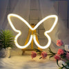 A glowing white neon sign shaped like a butterfly rests on a wooden easel with pink roses in the foreground.