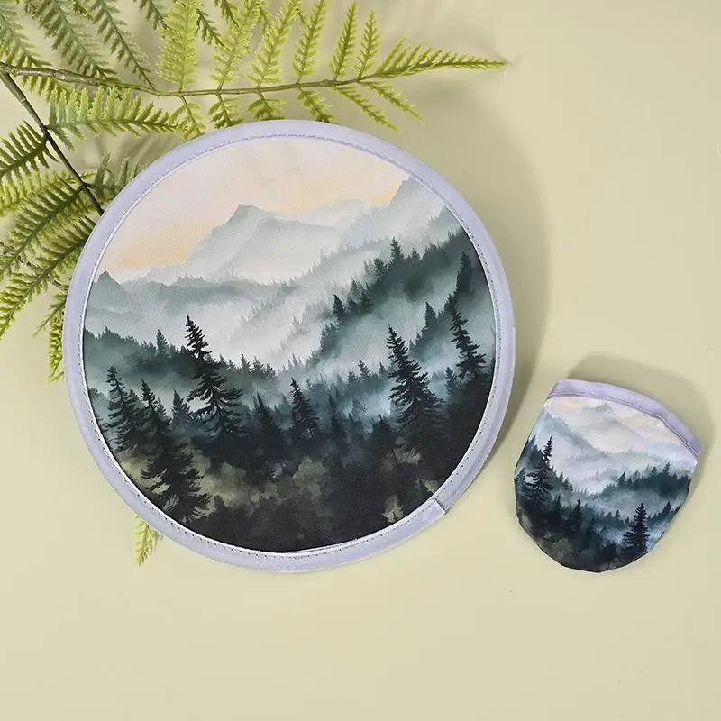 A round fabric fan featuring a misty forest landscape with evergreen trees and mountains.
