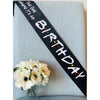 A black ribbon with white text reads Birthday where it's my on a light blue fabric background with white flowers.