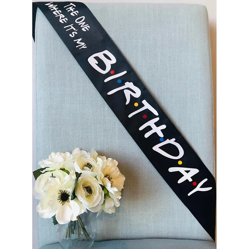 A black ribbon with white text reads Birthday where it's my on a light blue fabric background with white flowers.