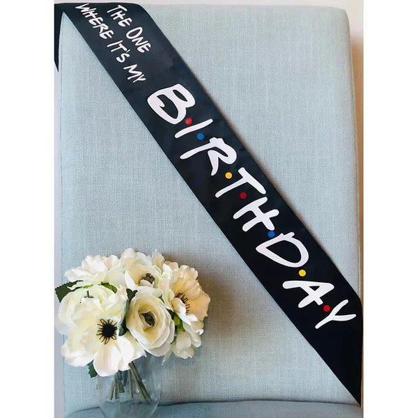 A black ribbon with white text reads Birthday where it's my on a light blue fabric background with white flowers.