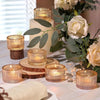 A collection of gold glass tealight candle holders in various sizes and wood slices arranged on a white table with greenery.