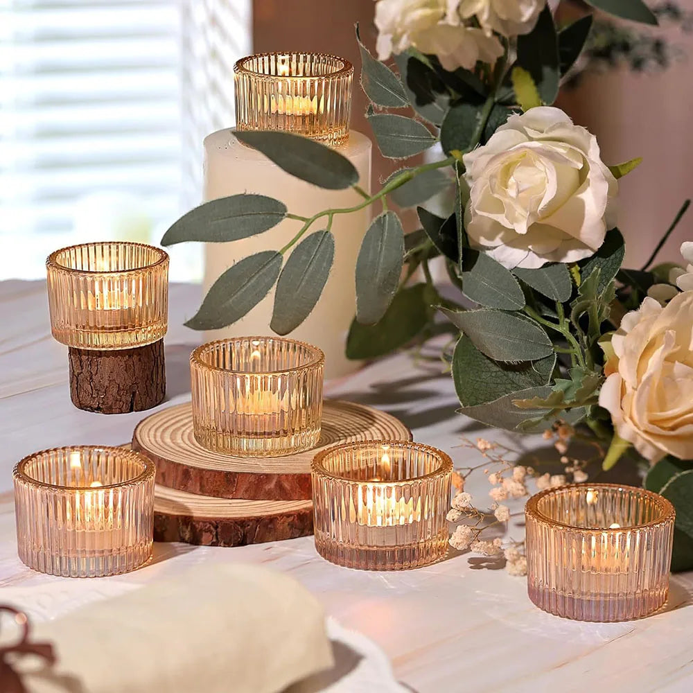 A collection of gold glass tealight candle holders in various sizes and wood slices arranged on a white table with greenery.