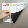A hand holds a magnetic fridge planner with monthly grids and erasable notes on a gray wall.