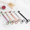 A collection of stainless steel candle wick trimmer scissors and a wooden tray on a white marble surface.