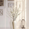 A branch of white faux plum blossoms with stems displayed in a light-colored vase.