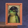 A framed picture of a green frog with decorative silver hair accessories.