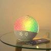 A sunrise alarm clock with a colorful, gradient light display sits on a glass table next to a smartphone.