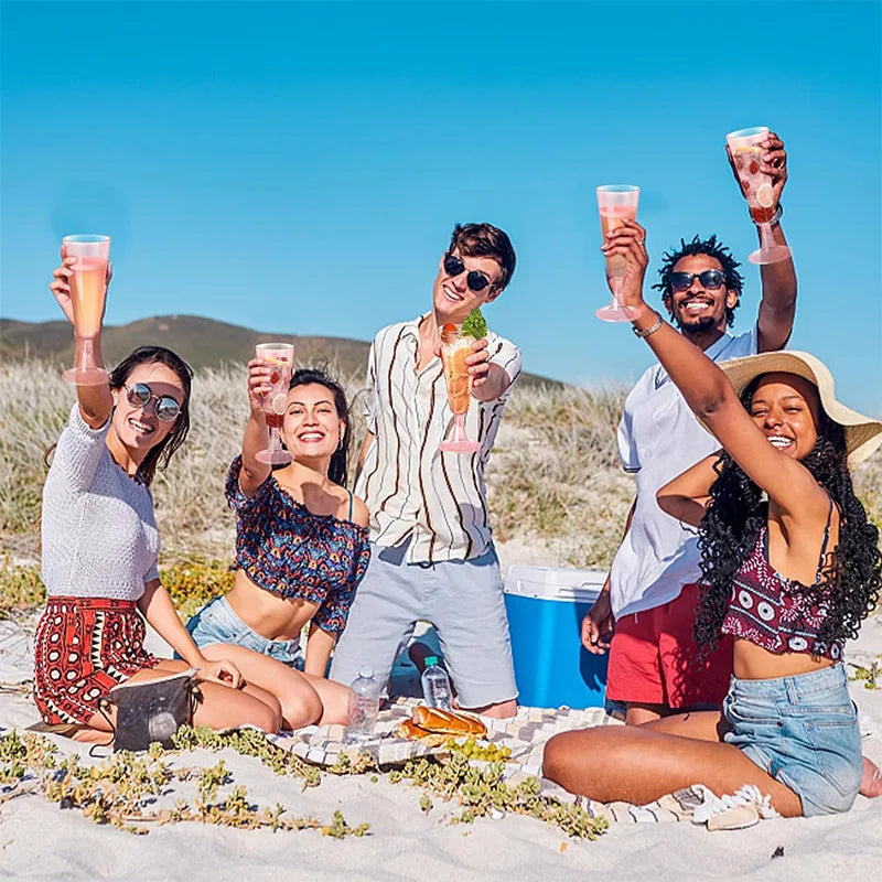 Five people celebrating outdoors with elegant disposable champagne glasses on a sandy beach under a blue sky