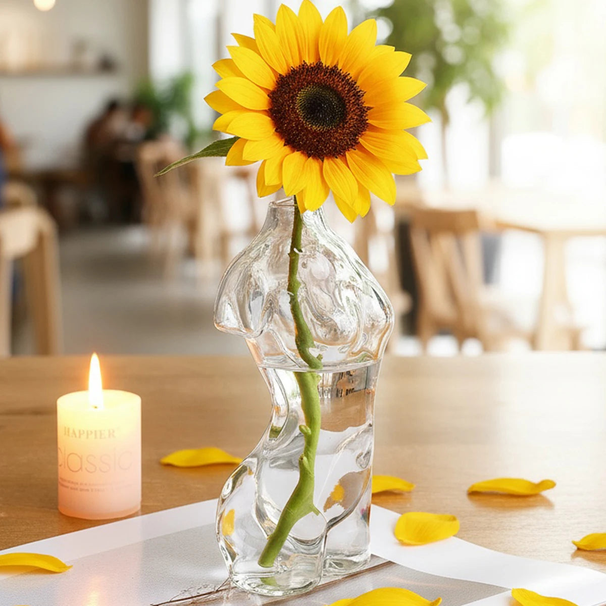 Artistic glass human body vase holding a single yellow sunflower in a bright indoor setting.