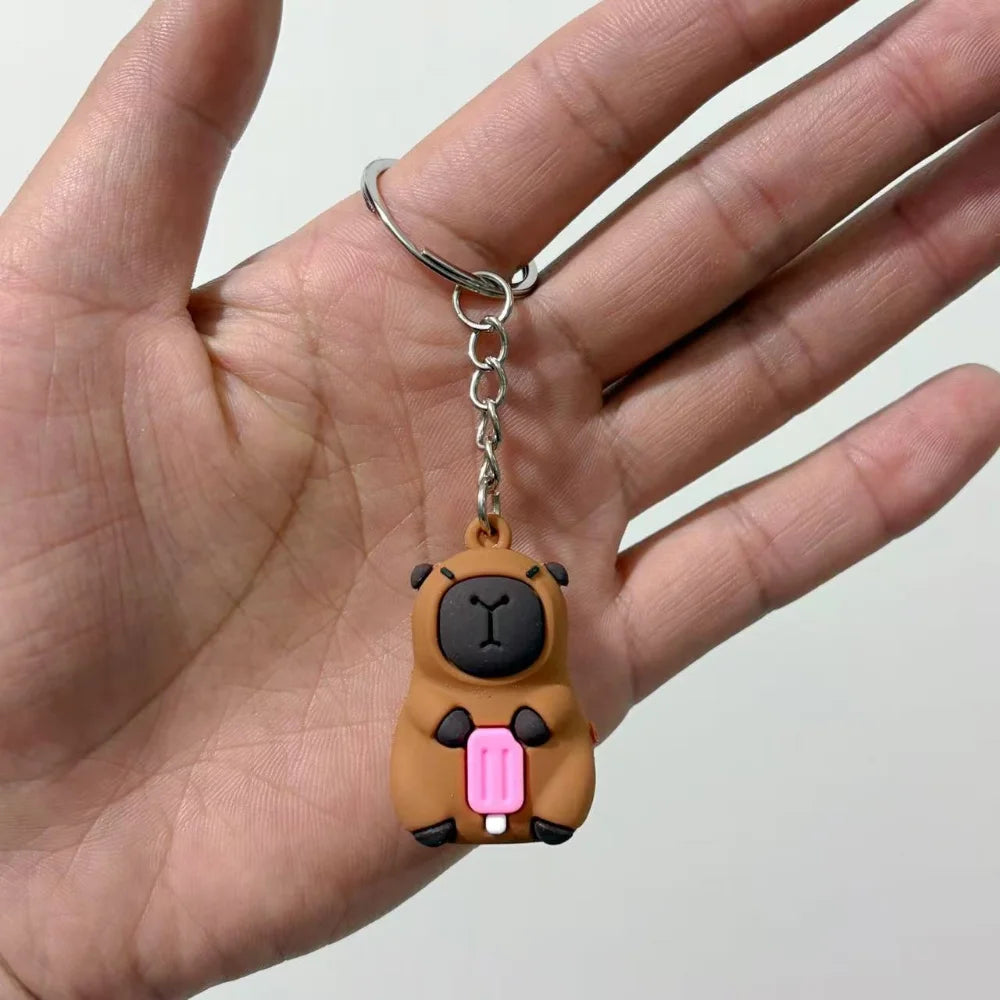 Un petit pendentif de clé en forme de capybara brun avec des accents roses est tenu dans la main d'une personne.