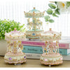 A set of delicate pink and gold resin carousel music boxes displayed on a wooden surface with greenery and books.