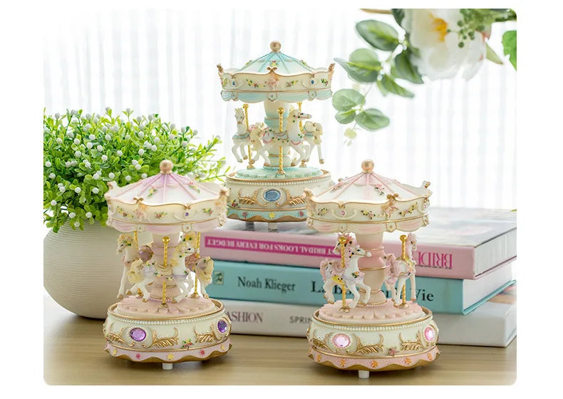 A set of delicate pink and gold resin carousel music boxes displayed on a wooden surface with greenery and books.