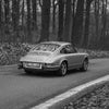 Une Porsche 911 argentée roule sur une route entourée d'une forêt dense en noir et blanc.