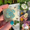A hand holds a small gold metal flower press kit featuring a turquoise flower, with dried yellow flowers and green foliage in
