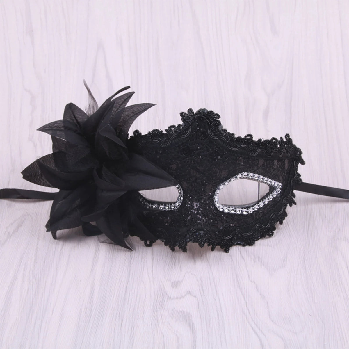 A black lace masquerade mask adorned with rhinestones and a large black feather is displayed on a white wooden surface.