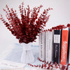 A bouquet of deep red artificial eucalyptus leaves displayed in a white vase on a light blue cloth.