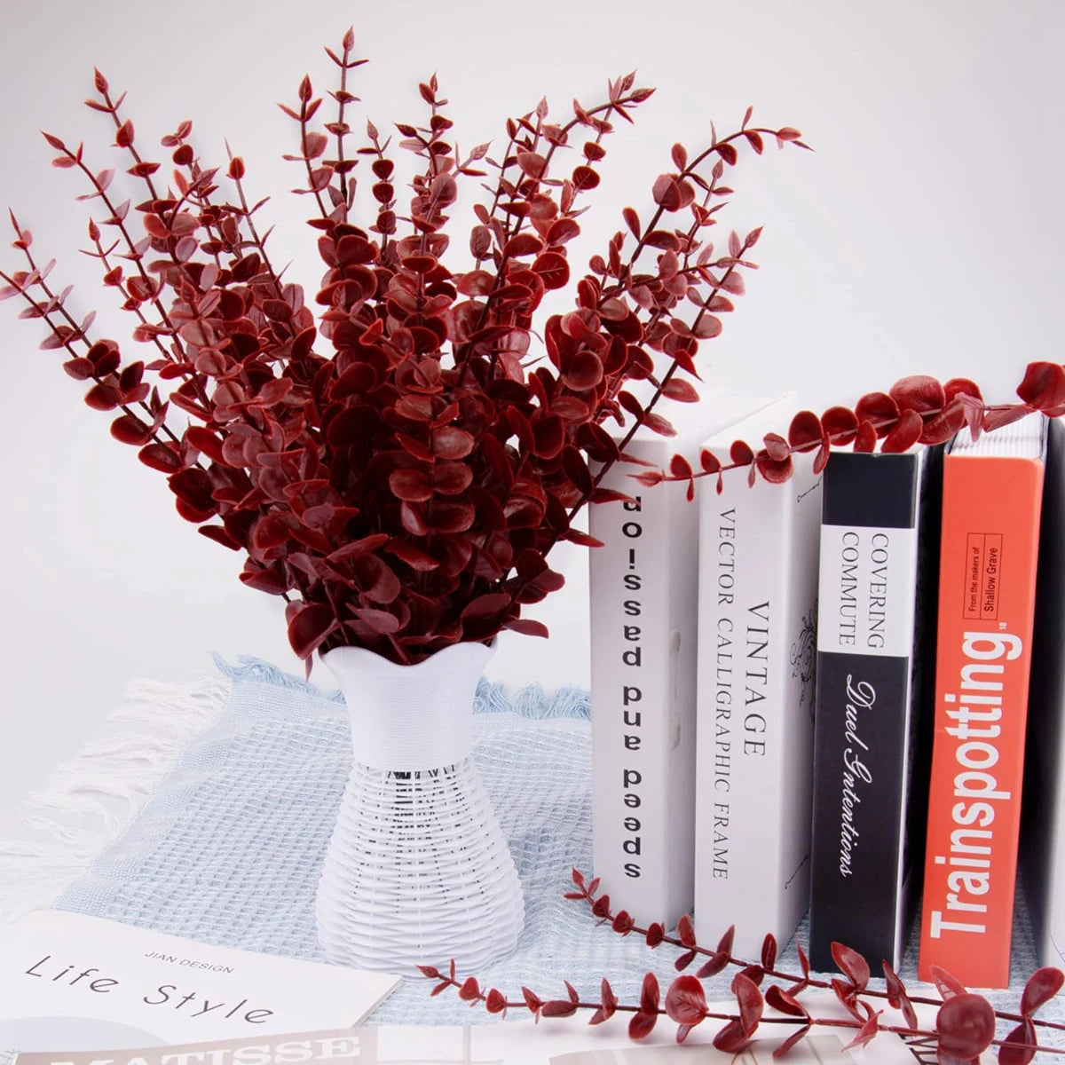 A bouquet of deep red artificial eucalyptus leaves displayed in a white vase on a light blue cloth.