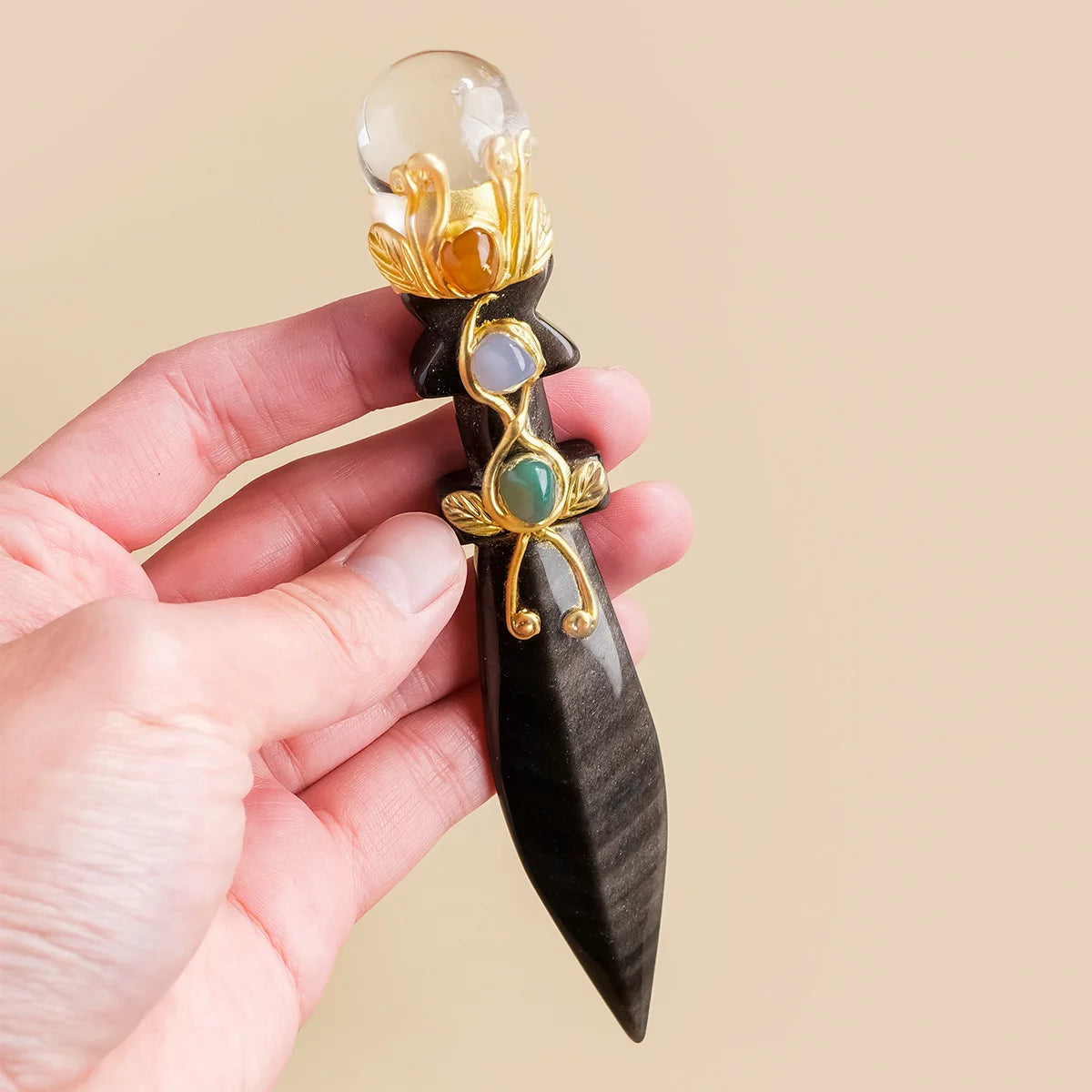 A hand holds a decorative crystal ball featuring amethyst and obsidian stones set in gold-toned metalwork on a dark wood piec