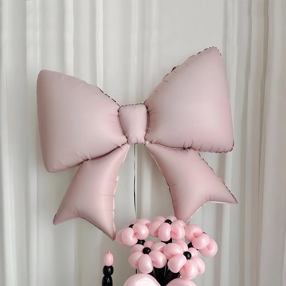 A pink bow-shaped foil balloon is displayed against a white curtain backdrop.