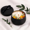 A black candle jar holds a white candle with a lit wick surrounded by dried floral elements on a white cloth.
