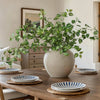 A large potted artificial eucalyptus stem with silk green leaves sits on a wooden table.
