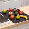 A black silicone drain mat is placed on a wooden countertop, holding various fresh fruits like apples, grapes, and bananas.
