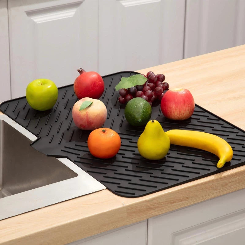 A black silicone drain mat is placed on a wooden countertop, holding various fresh fruits like apples, grapes, and bananas.