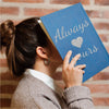 A person holds a blue notebook with silver glitter text that reads Always Yours against a brick background.