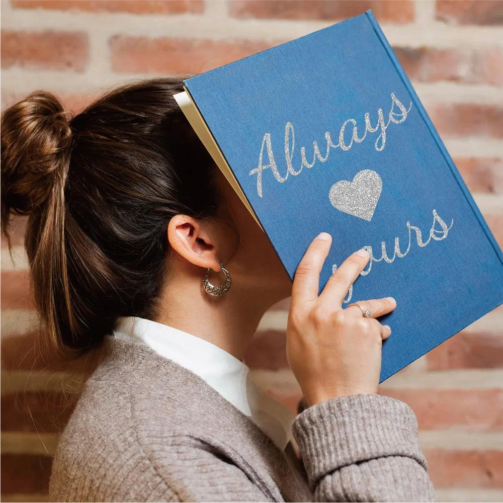 A person holds a blue notebook with silver glitter text that reads Always Yours against a brick background.