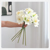 A hand holds a bouquet of white artificial clivia amaryllis flowers against a white background.