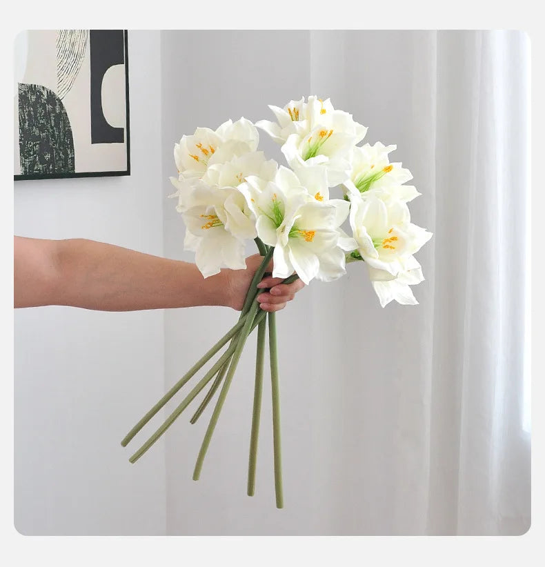 A hand holds a bouquet of white artificial clivia amaryllis flowers against a white background.