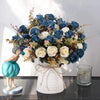 A bouquet of realistic silk blue and white roses arranged in a white vase on a dark surface.