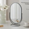 A silver oval vanity mirror with a rotating base sits on a white table next to a white flower and a cup.