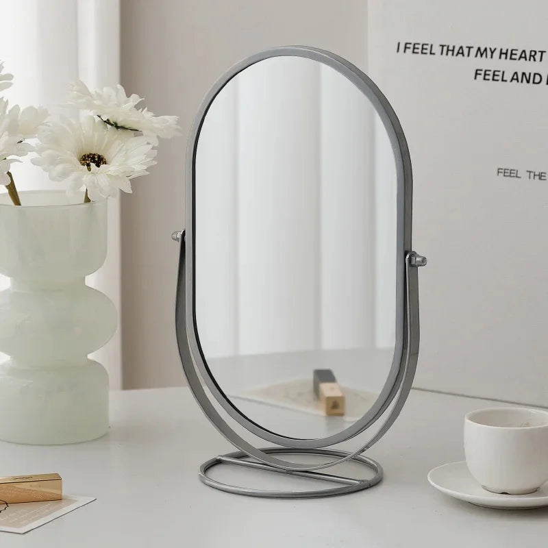 A silver oval vanity mirror with a rotating base sits on a white table next to a white flower and a cup.