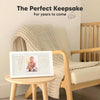 A white keepsake frame showing a baby's handprint and footprint, resting on a wooden table next to a knitted chair.